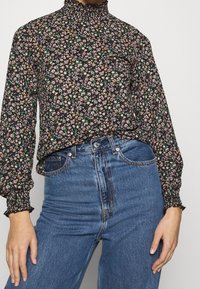 Zwarte bloes met bloemenprint, lange, geplooide mouwen en een hoge halslijn, met meerkleurige bloemenpatronen op zachte stof. Gecombineerd met blauwe jeans.