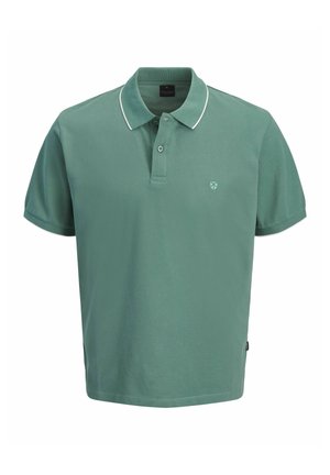 Szőke hajú fiú, zöld pólót, farmer rövidnadrágot, fehér zoknit és bézs cipőt visel, áll és oldalra néz.