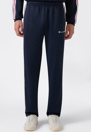 Uomo con pantaloni della tuta Champion blu navy con vita elastica e scarpe da ginnastica bianche su uno sfondo chiaro uniforme.