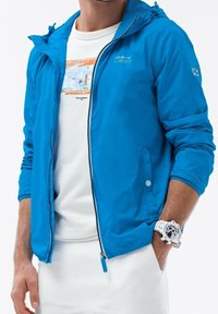 Blå letvægtsjakke med lynlås og hætte, med elastiske manchetter og trykknap. Båret over en hvid t-shirt.