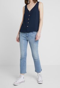 Mörkblå ärmlös topp med V-ringning och fem guldknappar, i kombination med ljusblå jeans och vita högskurna sneakers. Tydlig tygstruktur.