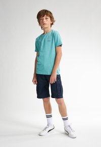 Tyrkysové tričko s krátkým rukávem a kulatým výstřihem, námořnické khaki šortky, bílé tenisky a pruhované černo-bílé ponožky. Jednoduchý design.