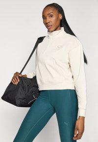Pull en polaire crème avec un col zippé moitié, poche avant et détail du logo ; associé à des leggings teal et un sac de sport noir.