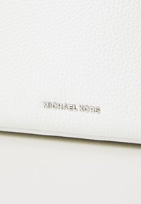 Vitt läderhandtag med en texturerad yta, med en silverfärgad metallisk "MICHAEL KORS"-logotyp präglad på framsidan.