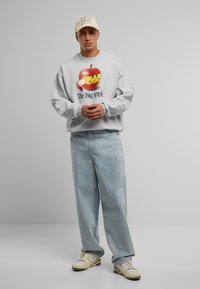 Grå sweatshirt med æblegrafik og teksten "The Big Apple", parret med lyseblå jeans og beige sneakers. Ensfarvet baggrund.