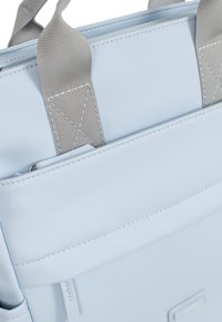 Tote bag en simili cuir bleu clair avec des poignées en tissu gris. Présente une poche avant et des détails de couture blanche le long des coutures.