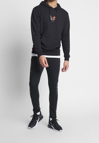 Felpa nera con grafica di farfalla sul petto, tasca a canguro, indossata sopra una maglietta bianca, abbinata a jeans skinny neri e scarpe da ginnastica nere.