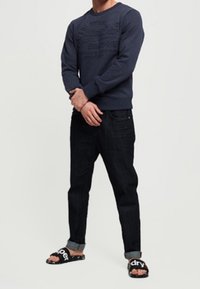 Felpa navy con branding in rilievo, jeans scuri e sandali neri con testo bianco. Vestibilità casual con orli arrotolati sui jeans.