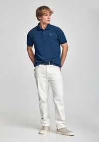 Navyblaues Poloshirt mit drei Knöpfen und gesticktem Logo, kombiniert mit weißen Jeans und beigen Sneakers. Sanfte Textur, lässige Passform.