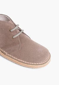 Scarpe in suede marrone con punta tondeggiante, lacci grigi e cuciture chiare lungo il bordo. Presentano una suola in gomma naturale e una fodera interna liscia.