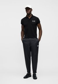 Homme portant un polo EA7 noir, un pantalon texturé noir, des baskets noires et une casquette blanche, debout les mains dans les poches.