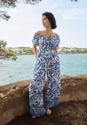 Žena stojí u kamenné zdi poblíž moře, má na sobě modrý a bílý květovaný overal s vykrojenými rameny, širokými nohavicemi a sandály.