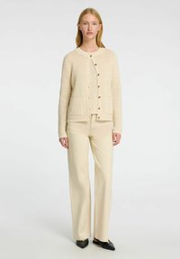 Béžový pletený kardigan s kulatým výstřihem, předním zapínáním na knoflíky a detaily kapes, zkombinovaný s béžovými kalhotami s širokými nohavicemi a černými botami.