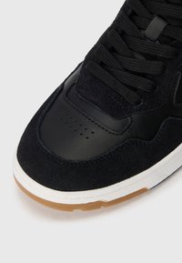 Black sneaker with suede and leather upper, flat laces, perforated toe area, and gum rubber outsole. Smooth, textured surfaces with detailed stitching.