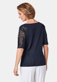 Marineblauer Pullover mit kurzen Ärmeln, der auf den Schultern mit aufwendiger schwarzer Spitzenverzierung und dezenten Verzierungen versehen ist. Glatte Strickstruktur.