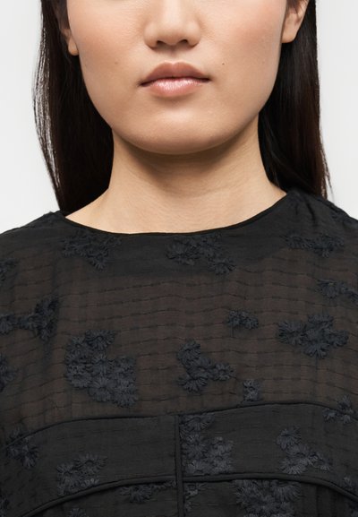 Κοντινό πλάνο του κάτω μέρους του προσώπου και του λαιμού μιας γυναίκας που φοράει μαύρο υφασμάτινο τοπ με κεντήματα λουλουδιών.