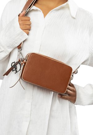 Borsa a tracolla marrone con texture, hardware argentato e tracolla in pelle, con frange decorative e un charm con logo, tenuta contro una camicia bianca.