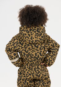 Barn med krøllet hår iført en brun og sort leopardmønstret hættejakke, med hænderne på hoften, vendt væk.