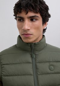 Young man with dark hair wearing a zipped-up olive green quilted jacket with round logo patch on a plain background.