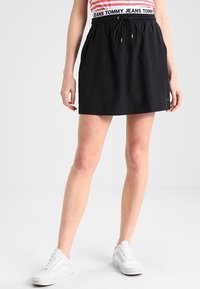 Svart idrottskjol med elastisk midja och dragsko. Slät tyg, knälång, ihop med vita sneakers.