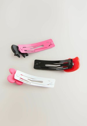 Trois accessoires capillaires colorés sont disposés sur un fond clair : une pince noire avec un accent rouge, une pince rose et une pince blanche avec un motif rose.
