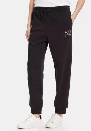 Pantaloni jogger neri con vita elastica e coulisse, con logo EA7 Emporio Armani sulla coscia, indossati con scarpe da ginnastica bianche.