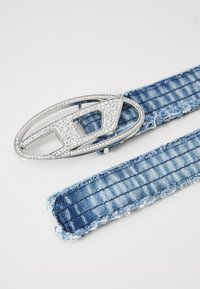 Denimbelte med frynser; blått stoff med vertikal søm. Sølv rektangulært spenne prydet med klare rhinestones og geometriske utskjæringer.