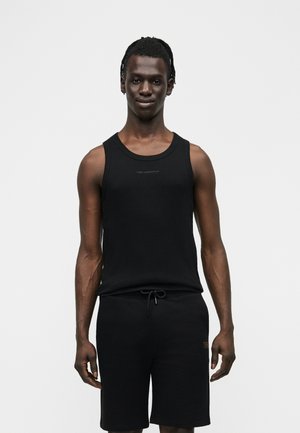 Giovane uomo con capelli intrecciati che indossa un top nero senza maniche e pantaloncini neri con coulisse, in piedi su uno sfondo bianco uniforme.