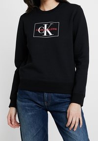 Czarna bluza z bawełny, z białym prostokątnym logo z napisem "Calvin Klein Jeans" w czerwonym kolorze, okrągły dekolt i ściągacze przy mankietach.
