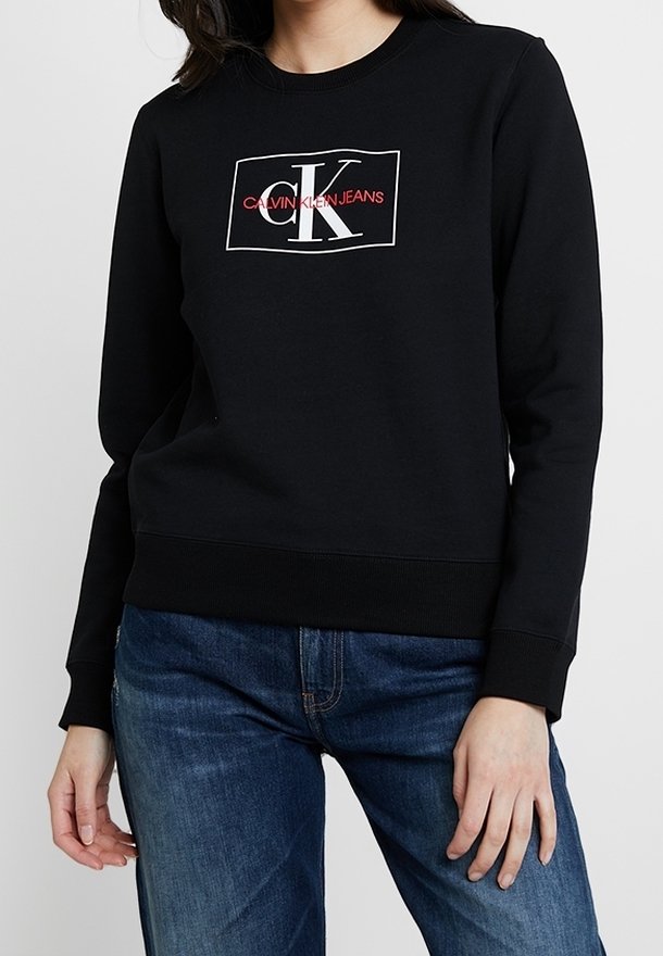 Czarna bluza z bawełny, z białym prostokątnym logo z napisem "Calvin Klein Jeans" w czerwonym kolorze, okrągły dekolt i ściągacze przy mankietach.