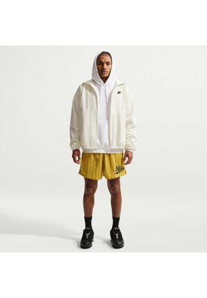 Mies seisoo kasvot eteenpäin, päällään valkoinen hupputakki, keltaiset Nike-shortsit, mustat sukat ja mustat lenkkarit yksiväristä taustaa vasten.