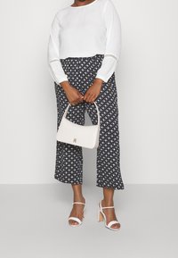 Maglietta bianca a maniche lunghe, pantaloni ampi a righe bianchi e neri, borsa leggera con logo, sandali bianchi con cinturini, texture del tessuto liscia.