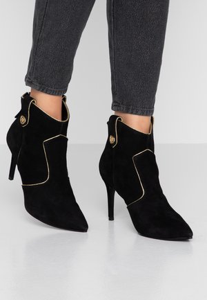 Zwarte suède enkellaarsjes met een spits toe, gouden bies en decoratieve knoopaccenten. De stilettohak zorgt voor extra hoogte en heeft een gladde textuur.