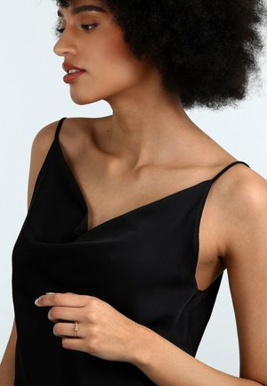 Schwarzes Satin-Cami-Top mit Spaghetti-Trägern und einem drapierten Ausschnitt. Eine Hand trägt einen goldenen Ring mit einem kleinen Edelstein am Finger.