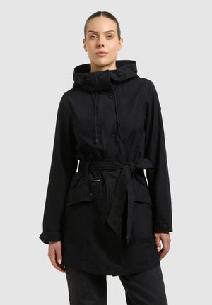 Kobieta w czarnej kurtce z kapturem i paskiem oraz dużymi przednimi kieszeniami, stojąca na tle jednolitego jasnoszarego tła.