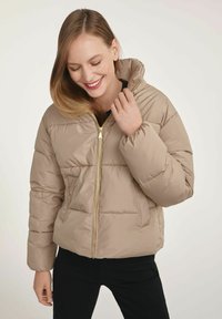 Kurtka puchowa w jasnym odcieniu beżu, z zamkiem z przodu, kapturem i pikowaną teksturą. Złote akcenty na suwakach oraz luźny krój z mankietami na nadgarstki.