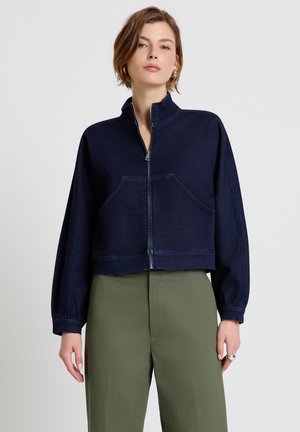 Femme portant une veste zippée bleu marine avec des poches avant et un pantalon taille haute vert olive sur un fond uni.