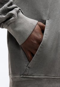 Χέρι σκουρόχρωμου δέρματος που έχει τοποθετηθεί στην μπροστινή τσέπη ενός γκρι φούτερ με ριμπ μανσέτα και τελείωμα, κοντινή λήψη.