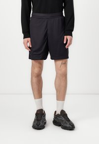 Shorts de sport noirs avec une texture lisse, taille élastique et poches latérales, associés à des baskets foncées et à des chaussettes blanches à mi-mollet.