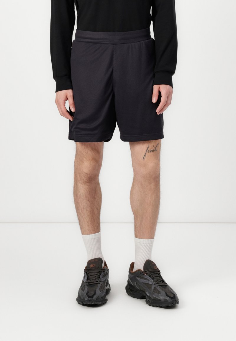 Shorts de sport noirs avec une texture lisse, taille élastique et poches latérales, associés à des baskets foncées et à des chaussettes blanches à mi-mollet.