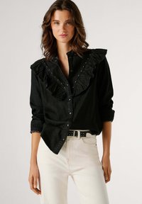Čierna rifľová košeľa s volánikmi na výstrihu, zapínaním na gombíky vpredu a striebornými cvokmi. Kombinovaná s krémovými džínsami a čiernym opaskom s bielymi bodkami.