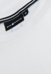 Bílý bavlněný tričko s kulatým výstřihem, s černou nálepkou a modrým nápisem, velikost "M". Hladká textura a jednoduchý design.