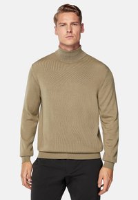 Knit turtleneck sweater in muted khaki, featuring a ribbed texture, long sleeves, and a relaxed fit, paired with black trousers.