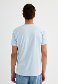 Camiseta de algodón azul claro con mangas cortas y escote redondo, llevada por una persona vista desde atrás, combinada con pantalones cortos de mezclilla clara.