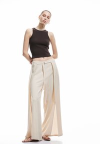 Top a maniche lunghe marrone a coste abbinato a pantaloni ampissimi color crema con fessure laterali, una superficie liscia e un dettaglio con zip centrale.