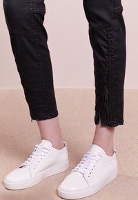 Pantalons noirs cropped avec des détails de couture visibles, dotés de fermetures éclair latérales aux poignets. Associés à des baskets blanches en cuir à semelles plates.