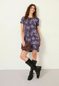 Kurzärmliges lila Blumenkleid mit figurbetonter Form, kombiniert mit knielangen schwarzen Stiefeln und einer kleinen bordeauxroten Handtasche. Glatte Stofftextur.