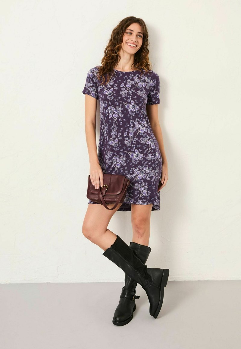 Kurzärmliges lila Blumenkleid mit figurbetonter Form, kombiniert mit knielangen schwarzen Stiefeln und einer kleinen bordeauxroten Handtasche. Glatte Stofftextur.