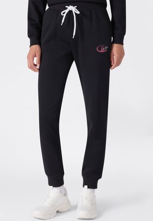 Person trägt schwarze Jogginghosen mit weißem Kordelzug, kleinem pink-weißem Logo am linken Oberschenkel, weißen Socken und weißen Turnschuhen.