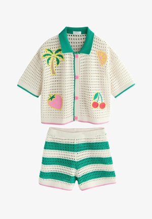 Ragazza con occhiali dalle lenti gialle, top all'uncinetto con toppe a forma di frutta e pantaloncini a righe all'uncinetto, in piedi davanti a uno sfondo beige in studio.